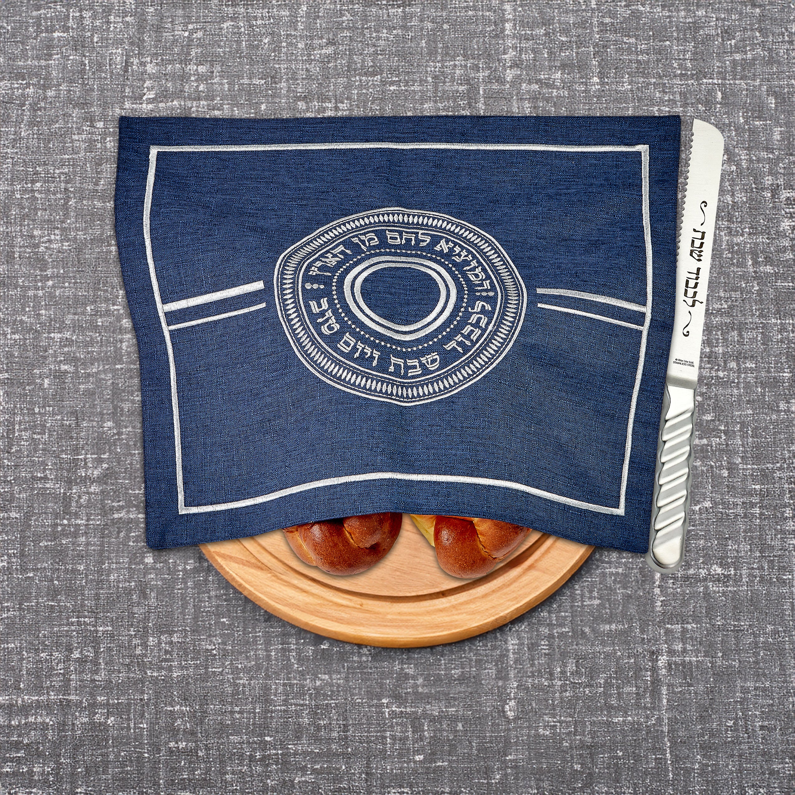 Blue challah cover with white design on a gray surface, featuring a wooden cutting board with bread and a knife.