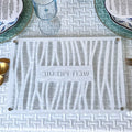 A challah cutting board with a geometric cutout pattern and the Hebrew text 'שבת יום טוב' (Shabbat Yom Tov) in the center, displayed on a white textured table runner with plates and glassware.