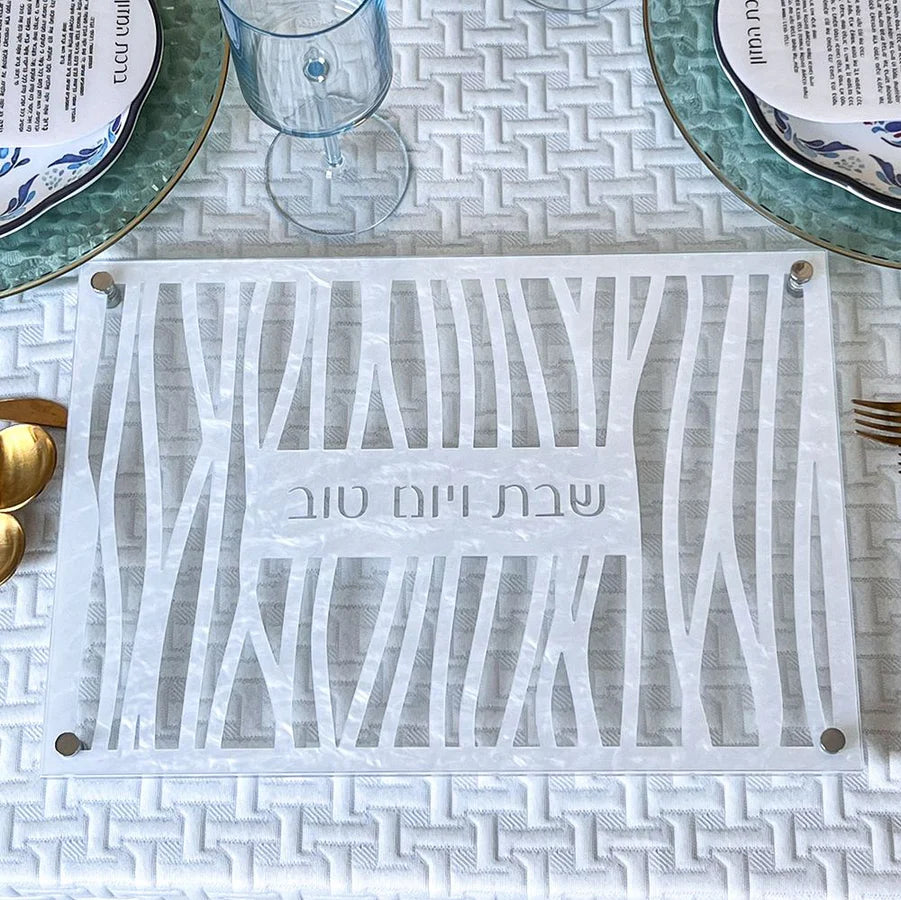 A challah cutting board with a geometric cutout pattern and the Hebrew text 'שבת יום טוב' (Shabbat Yom Tov) in the center, displayed on a white textured table runner with plates and glassware.