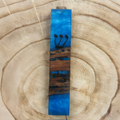A rectangular wooden mezuzah with a blue resin inlay and black Hebrew lettering, displayed on a natural wood slice background.