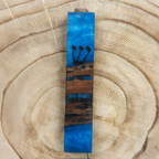 A rectangular wooden mezuzah with a blue resin inlay and black Hebrew lettering, displayed on a natural wood slice background.