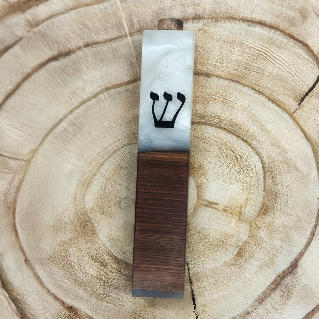 A mezuzah case made of white marble on the top half and dark brown wood grain on the bottom half, featuring a black Hebrew letter Shin, displayed on a circular wood slice background.