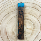 A tall, narrow wooden mezuzah case with a carved Hebrew letter 'Shin' and a small gold-toned ornament, featuring a blue and dark brown wood grain pattern, displayed on a light wood slice background.