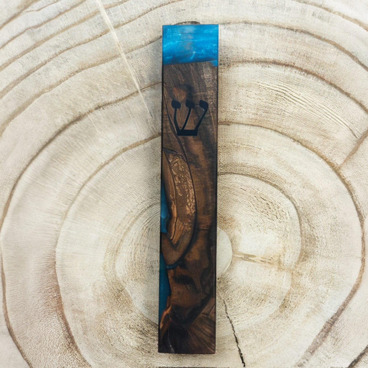 A tall, narrow wooden mezuzah case with a carved Hebrew letter 'Shin' and a small gold-toned ornament, featuring a blue and dark brown wood grain pattern, displayed on a light wood slice background.
