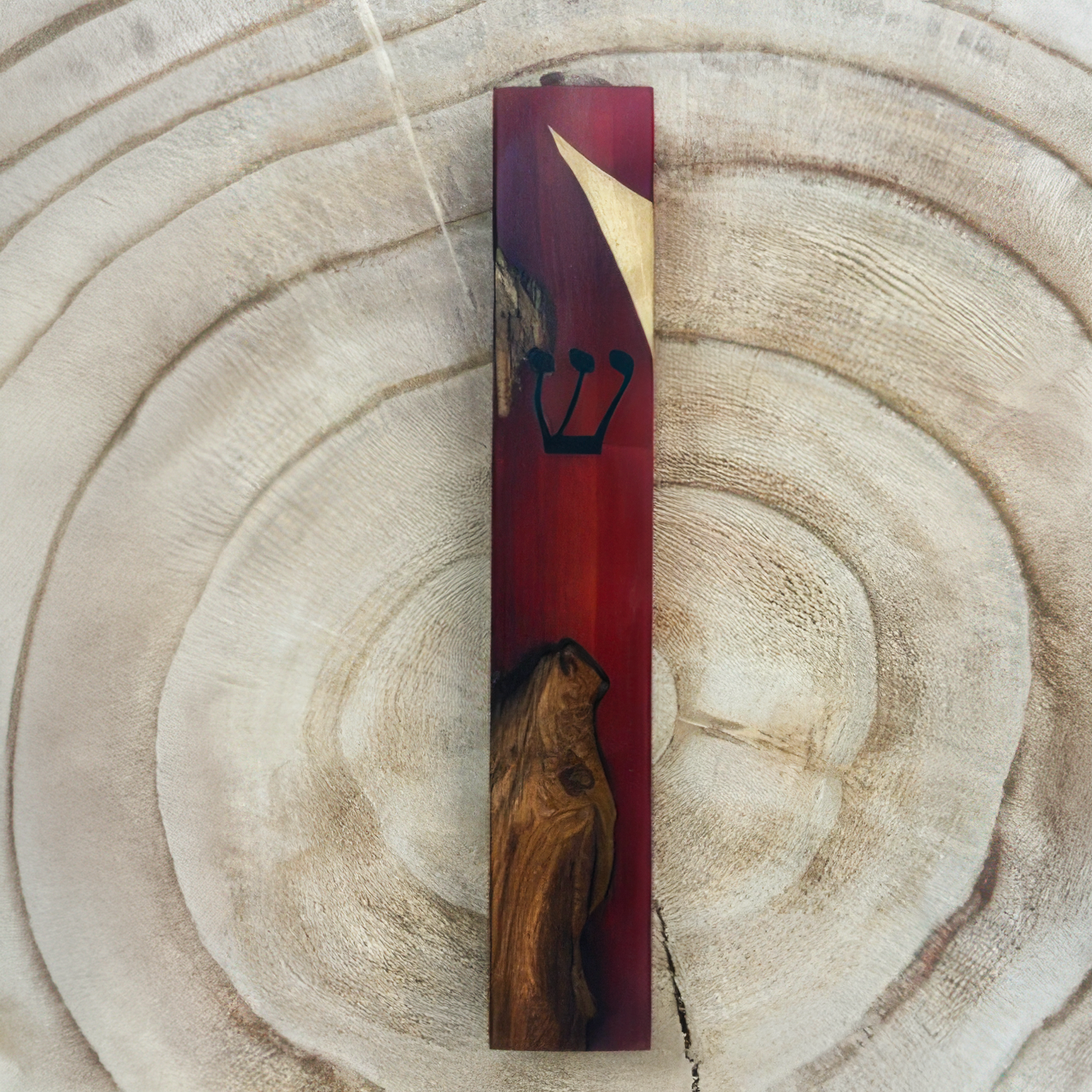 A rectangular wooden mezuzah case with a dark red resin body, featuring a black Hebrew letter 'Shin' and a gold-toned corner accent, displayed on a light wood grain background.