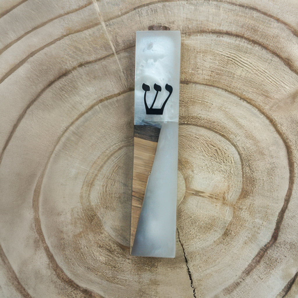 A rectangular mezuzah case made of white and gray marbled resin with a light wood inlay and a black Hebrew letter 'Shin', displayed on a natural wood slice background.