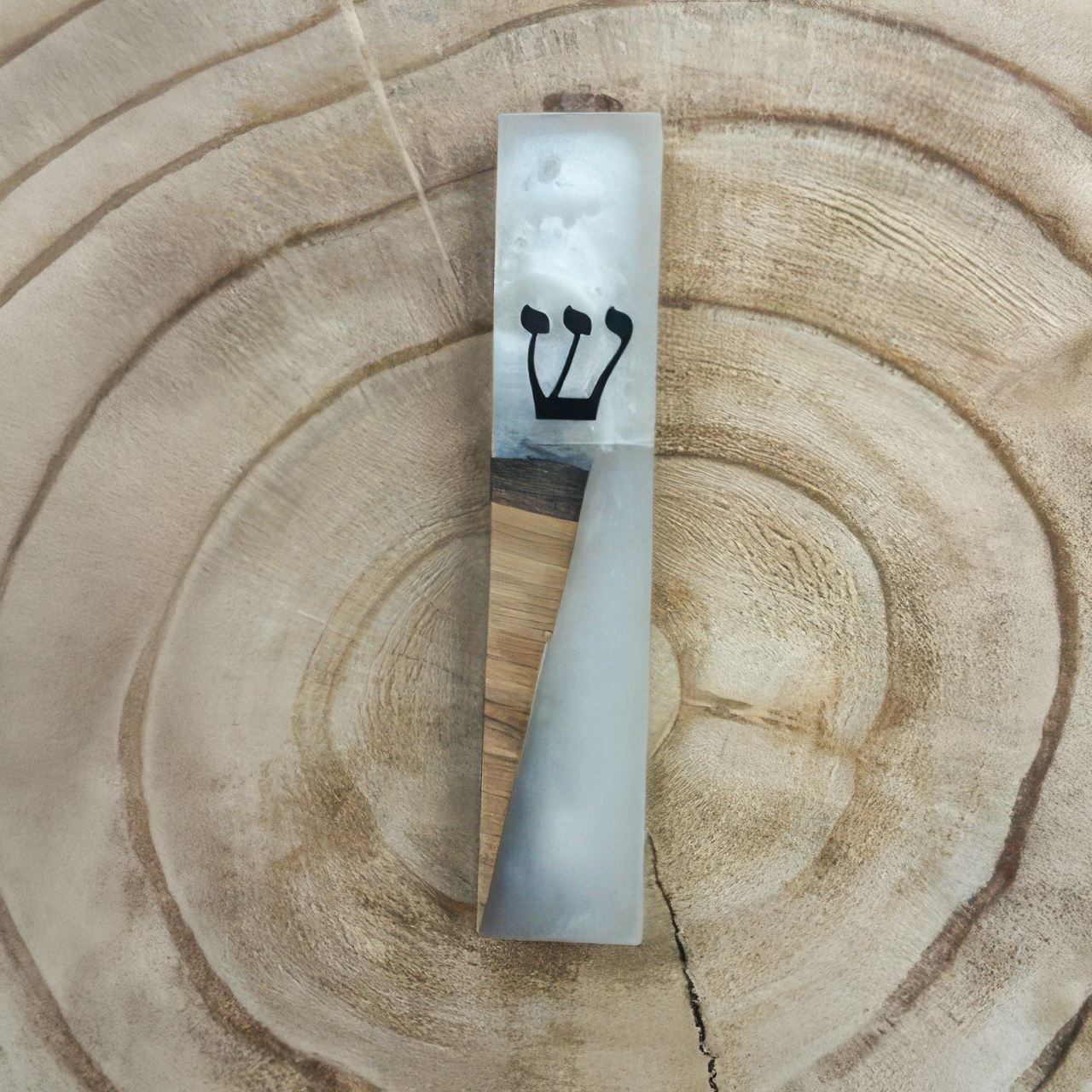 A rectangular mezuzah case made of white and gray marbled resin with a light wood inlay and a black Hebrew letter 'Shin', displayed on a natural wood slice background.