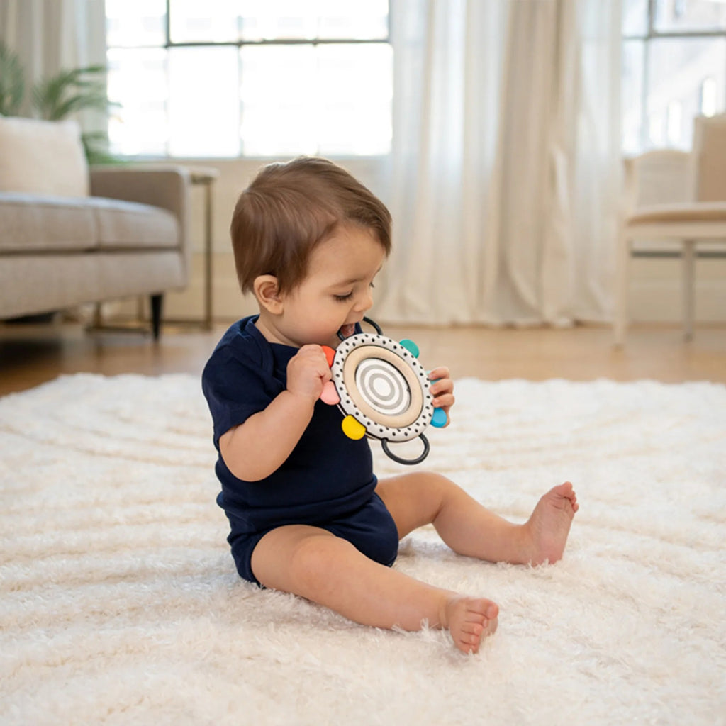 A baby sitting on a white shag rug in a bright living room, holding a circular sensory toy with colorful accents.