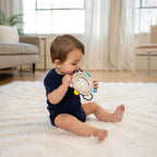 A baby sitting on a white shag rug in a bright living room, holding a circular sensory toy with colorful accents.