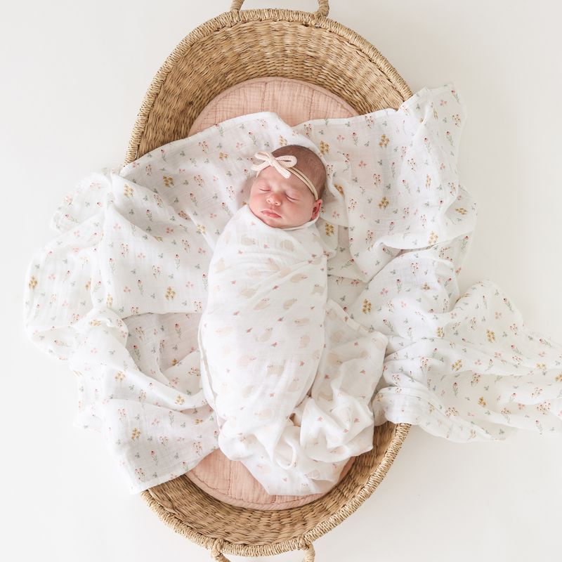 A sleeping newborn baby swaddled in a white muslin blanket with a tiny floral print, resting inside a woven oval basket on a soft pink cushion. The baby also wears a matching white bow headband.