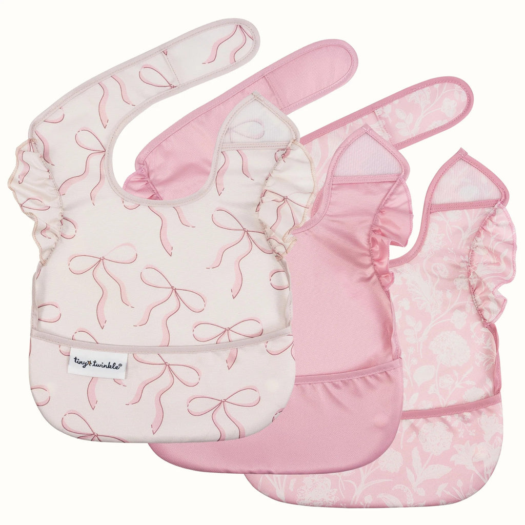 Three pink baby bibs with ruffled edges, overlapping on a light cream background. The bibs feature different prints: one with pink bow motifs on a white base, one solid light pink, and one with a pale pink floral pattern.