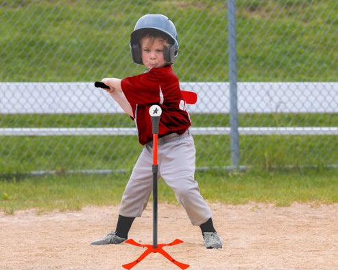 A young child wearing a gray helmet and a red-and-black baseball jersey stands on a baseball field, holding a T-ball bat on an orange T-stand. A chain-link fence and white bench are visible in the background.