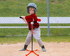 A young child wearing a gray helmet and a red-and-black baseball jersey stands on a baseball field, holding a T-ball bat on an orange T-stand. A chain-link fence and white bench are visible in the background.