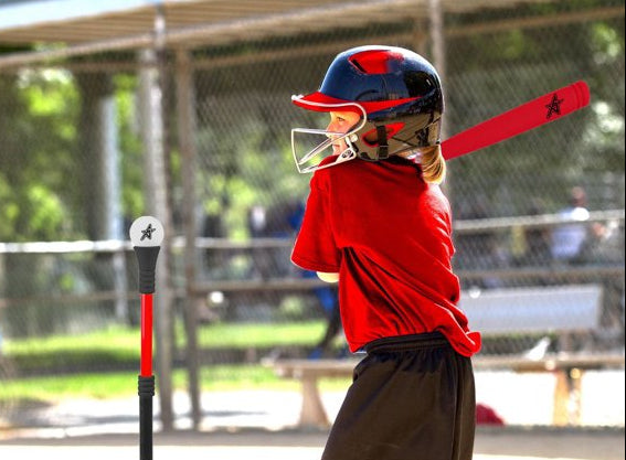A child wearing a red shirt, black shorts, and a red-and-black batting helmet holds a red bat near a T-ball training stand on a baseball field.