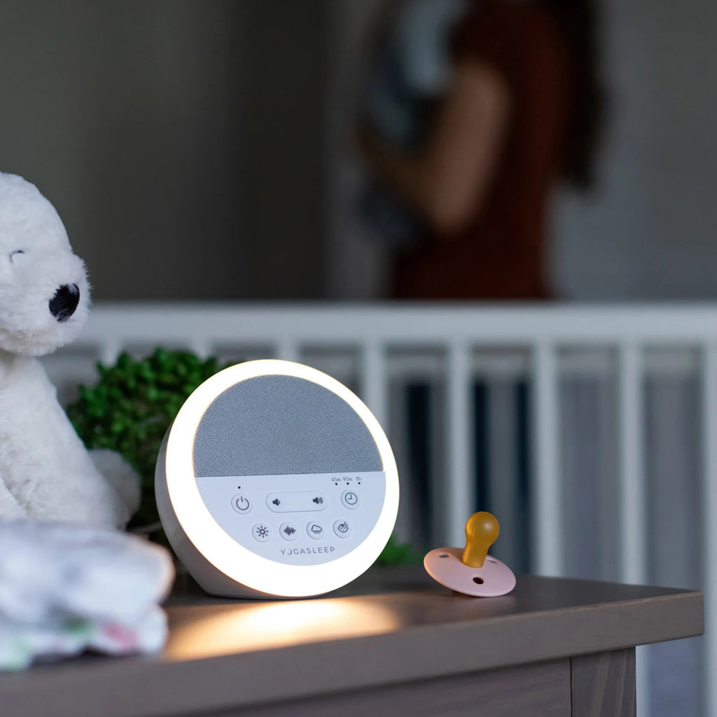 A round white baby sleep monitor with a glowing light ring sits on a bedside table next to a pacifier and a plush toy, with a blurred nursery background.