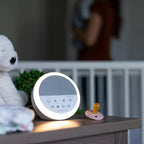 A round white baby sleep monitor with a glowing light ring sits on a bedside table next to a pacifier and a plush toy, with a blurred nursery background.