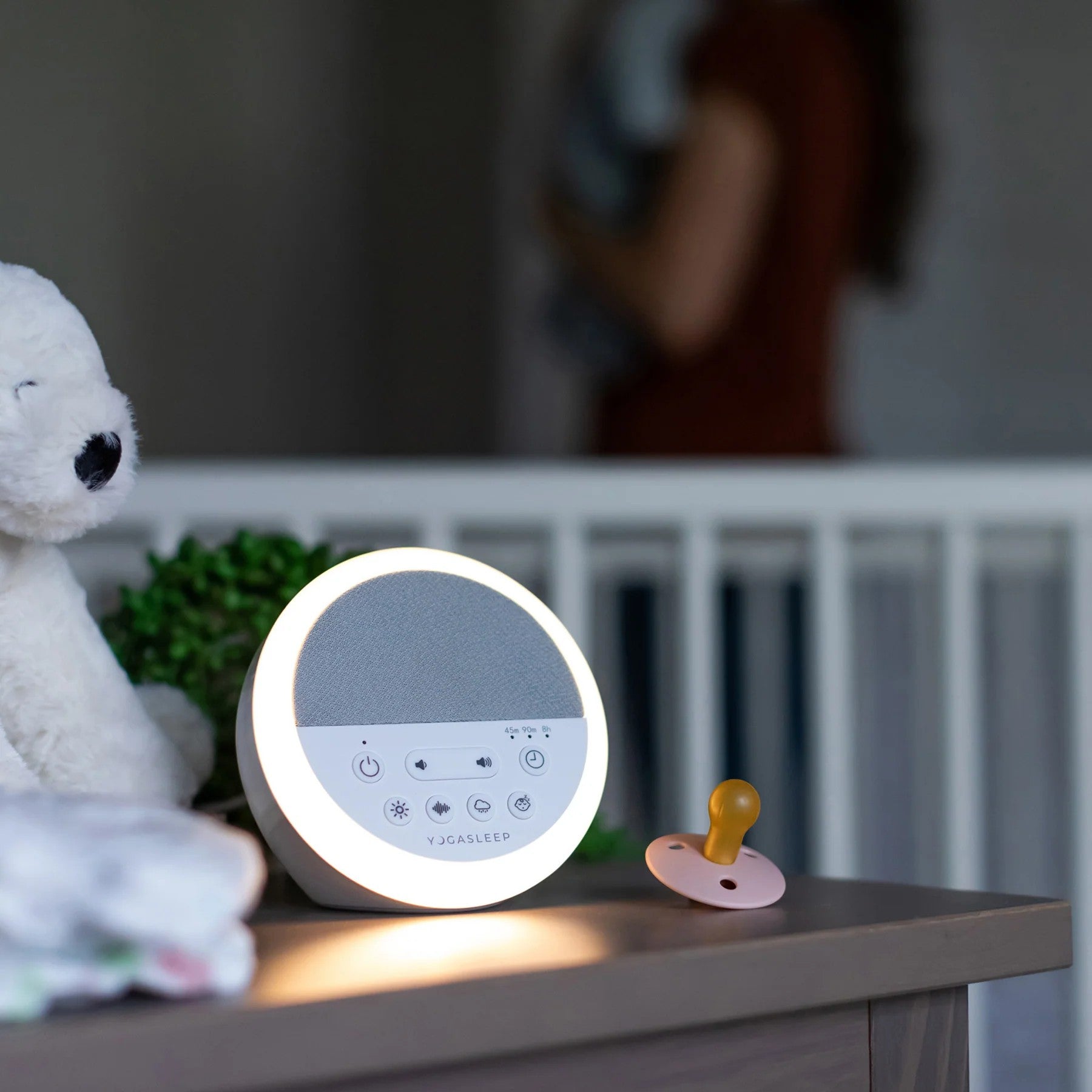 A round white baby sleep monitor with a glowing light ring sits on a bedside table next to a pacifier and a plush toy, with a blurred nursery background.