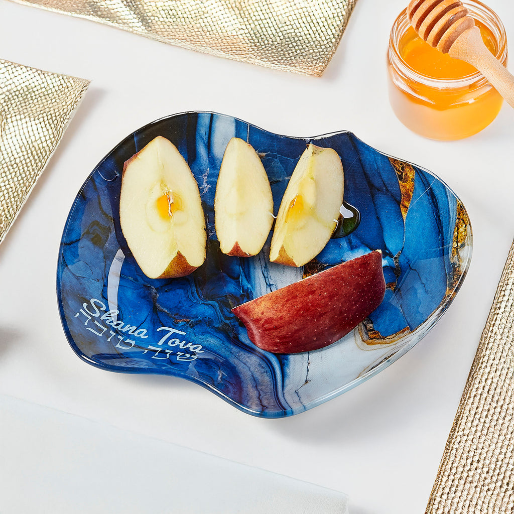 A blue and navy marbled glass appetizer plate with gold accents, styled with apple slices and a piece of red apple on a white surface with gold textured placemats and a jar of honey.