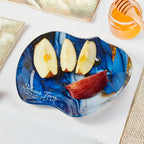A blue and navy marbled glass appetizer plate with gold accents, styled with apple slices and a piece of red apple on a white surface with gold textured placemats and a jar of honey.