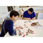 A woman and a man sit at a white table in a bright living room, assembling a colorful jigsaw puzzle. The puzzle box labeled 'Rosh Hashanah JIGSAW PUZZLE' and an open white box are on the table next to scattered puzzle pieces.