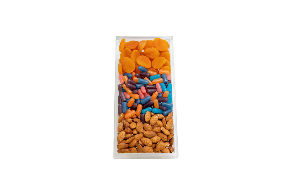 A clear plastic rectangular container filled with three distinct sections of dried fruits and nuts: orange apricots at the top, multicolored capsules in the middle, and almonds at the bottom, isolated on a white background.