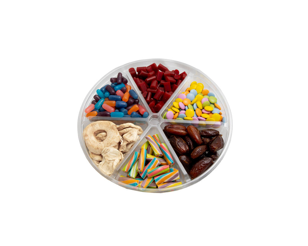 A clear plastic divided tray filled with assorted colorful candies, chocolate pieces, dried fruit slices, and rainbow-striped sticks, photographed on a plain white background.