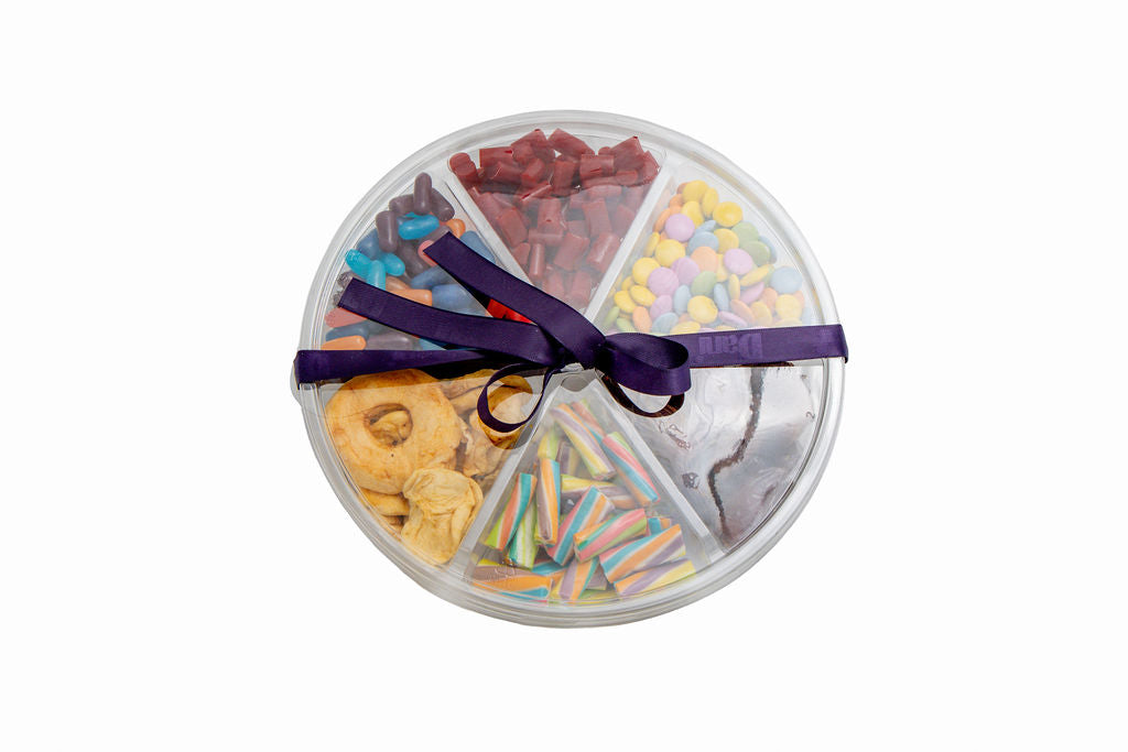 A round clear plastic gift box divided into six sections, filled with assorted colorful candies, chocolate pieces, and apples, tied with a dark purple ribbon, on a white background.