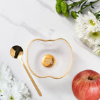Heart-shaped dish with gold rim and central emblem, accompanied by a gold spoon and an apple on a white background.