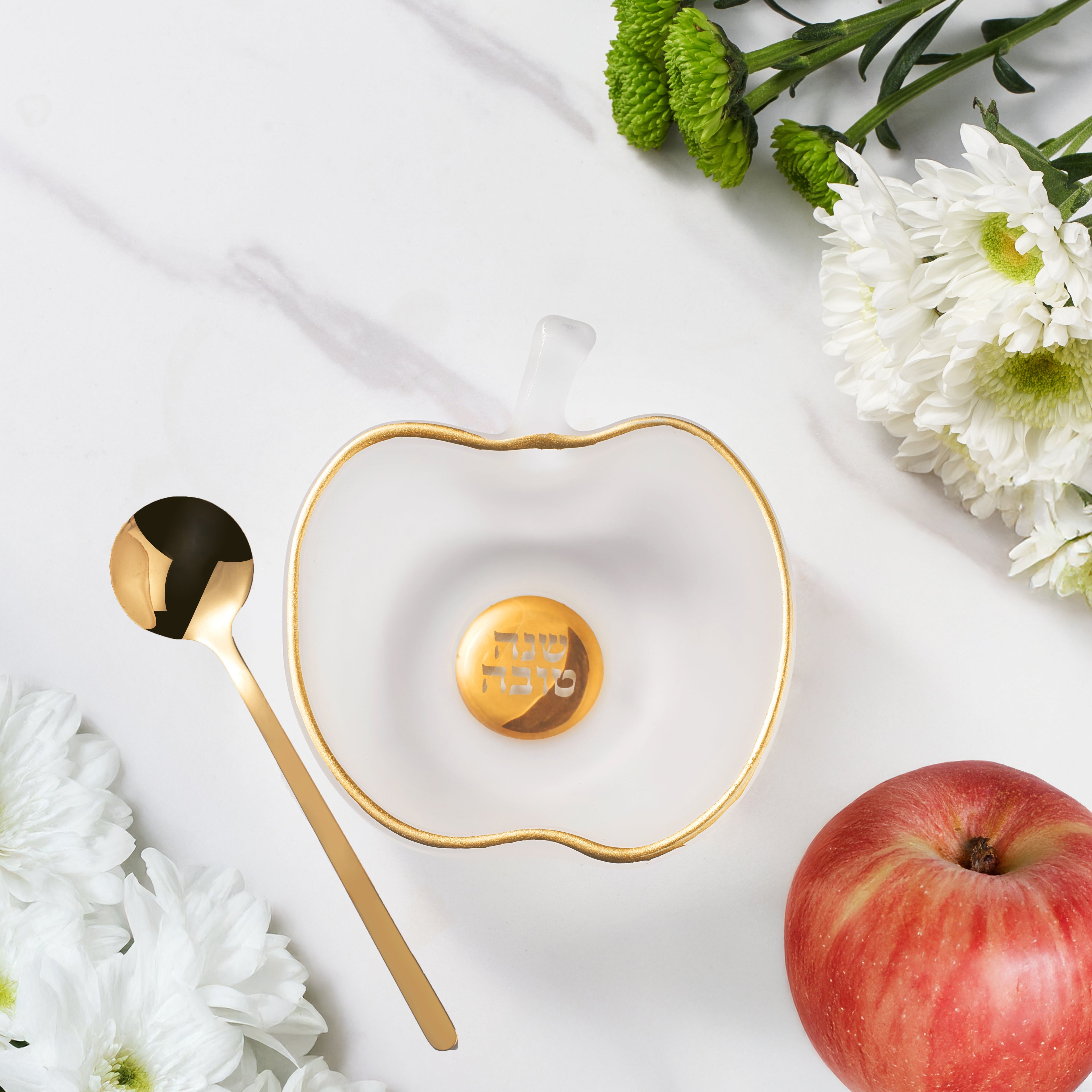 Heart-shaped dish with gold rim and central emblem, accompanied by a gold spoon and an apple on a white background.