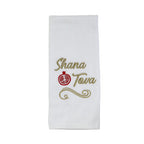 A white fabric tea towel with 'Shana Tova' embroidered in gold thread and a red pomegranate symbol in the center, displayed flat against a light off-white background.