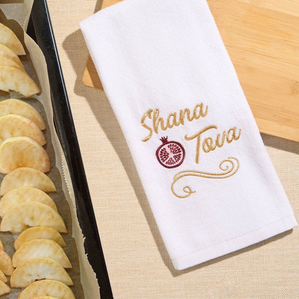 A white kitchen towel with gold embroidered text reading 'Shana Tova' and a pomegranate motif, laid on a beige surface beside a baking tray of apple slices.