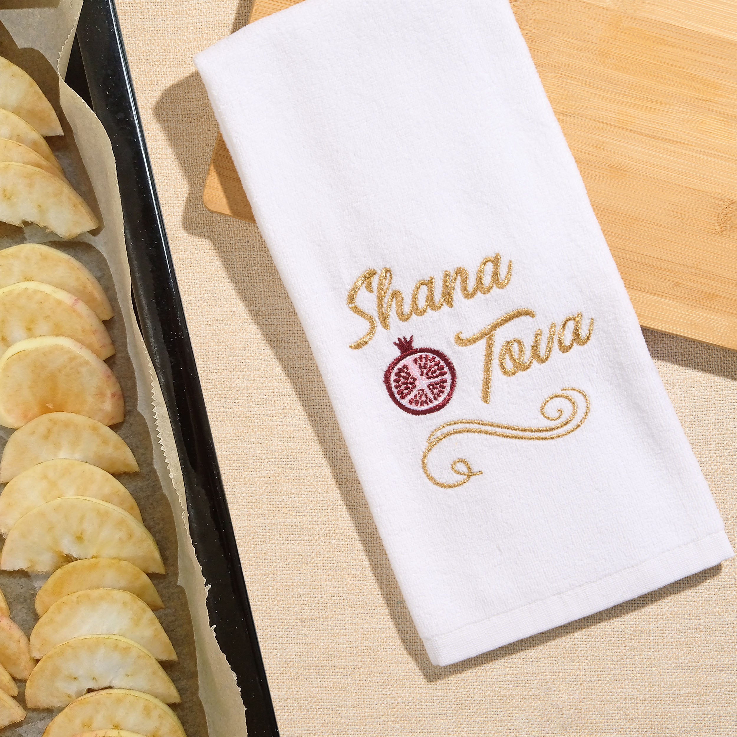 A white kitchen towel with gold embroidered text reading 'Shana Tova' and a pomegranate motif, laid on a beige surface beside a baking tray of apple slices.
