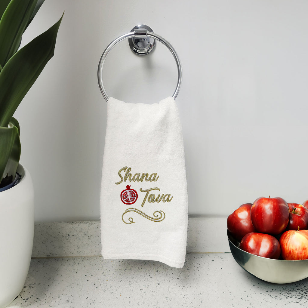A white cotton towel hanging from a silver wall-mounted ring holder on a bathroom counter. The towel features gold embroidered text reading 'Shana Tova' with a red pomegranate symbol in the center.