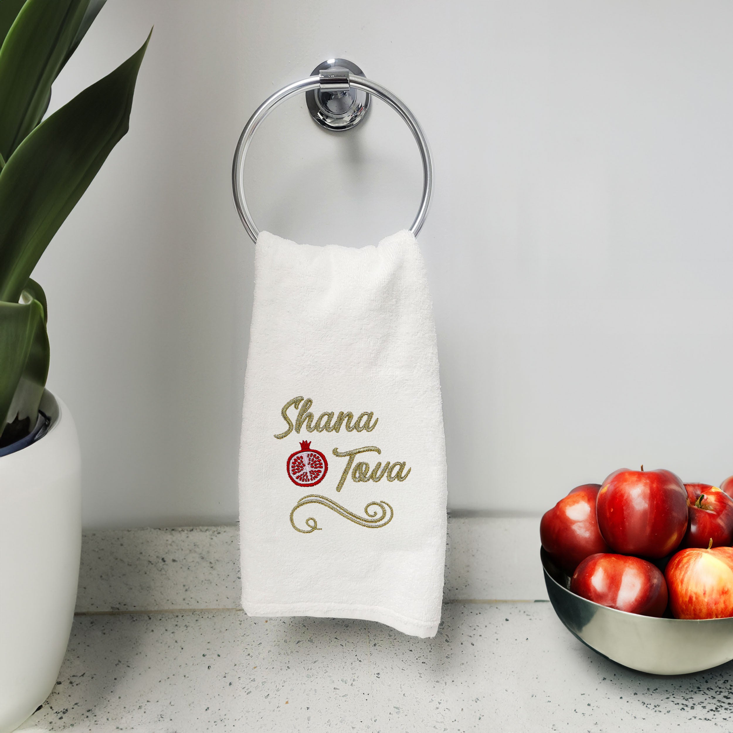 A white cotton towel hanging from a silver wall-mounted ring holder on a bathroom counter. The towel features gold embroidered text reading 'Shana Tova' with a red pomegranate symbol in the center.
