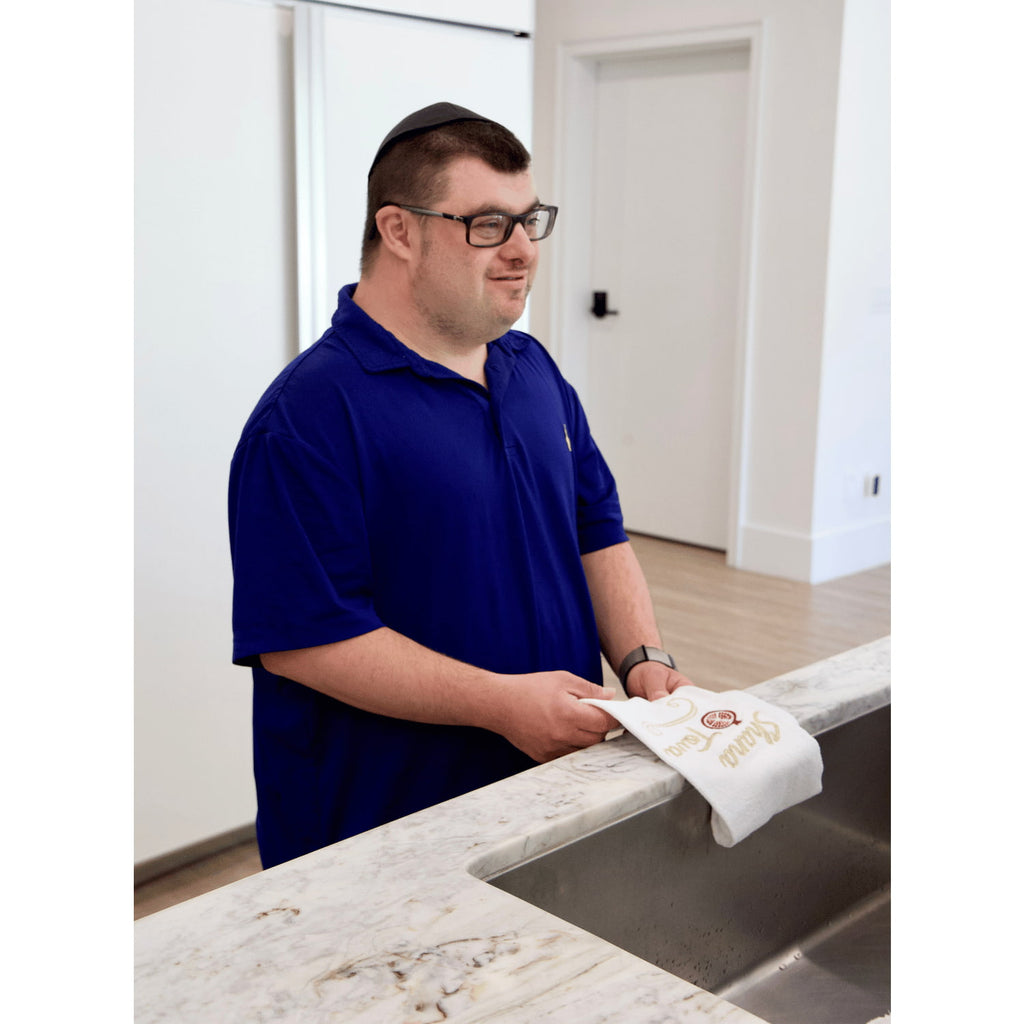 A man wearing a navy blue polo shirt and glasses stands in a kitchen, holding a white kitchen towel with gold embroidery featuring the word 'Shana Tova'.