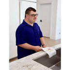 A man wearing a navy blue polo shirt and glasses stands in a kitchen, holding a white kitchen towel with gold embroidery featuring the word 'Shana Tova'.