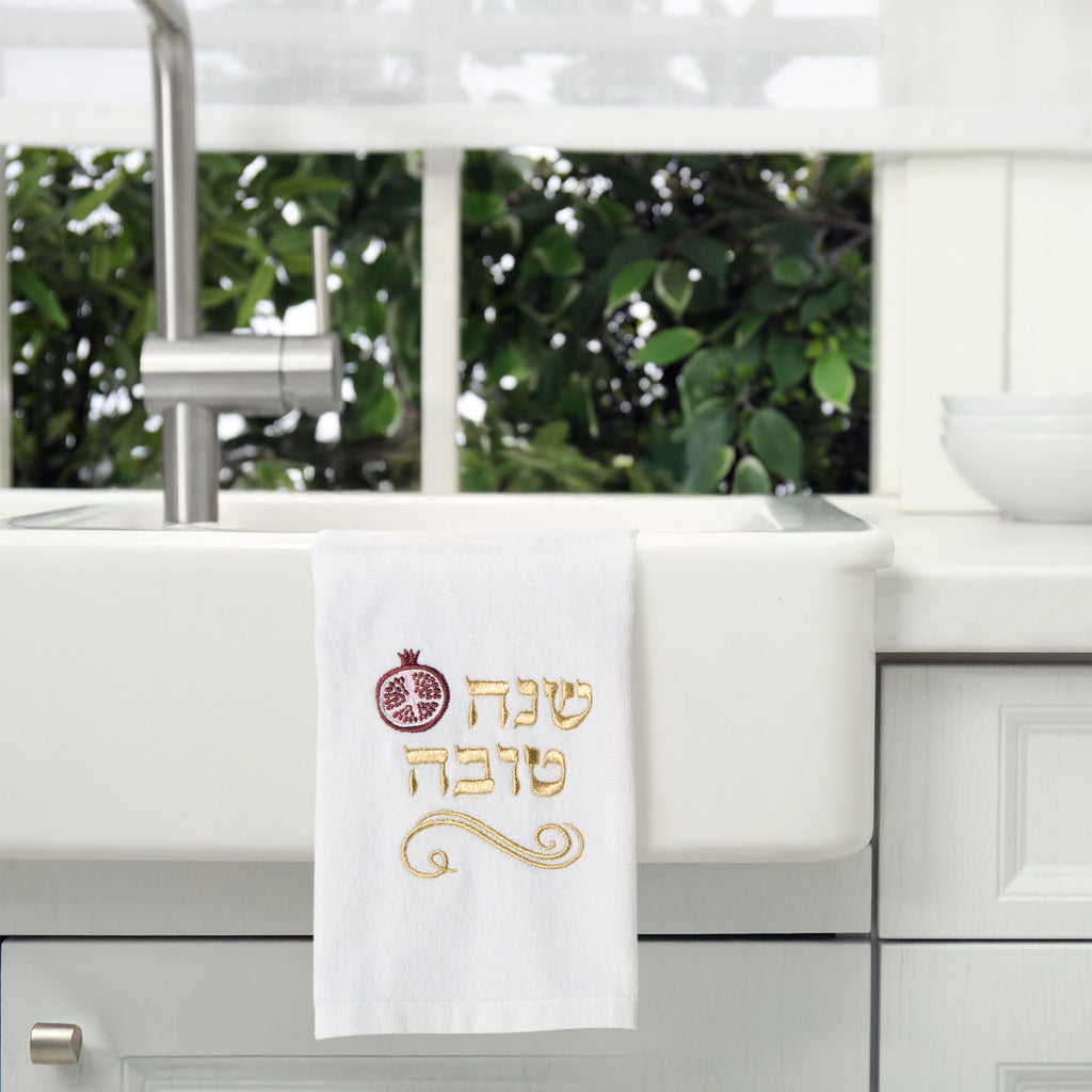 A white kitchen towel with gold and red embroidery hanging over a white farmhouse sink. The embroidery features a pomegranate symbol and Hebrew text reading 'Shenah Tova' in gold with a red pomegranate accent.