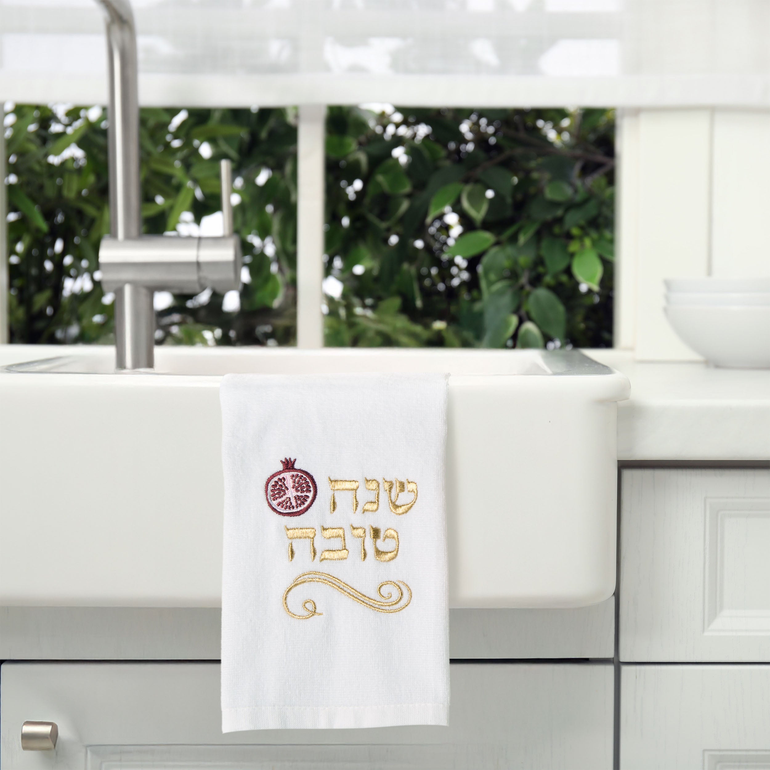 A white kitchen towel with gold and red embroidery hanging over a white farmhouse sink. The embroidery features a pomegranate symbol and Hebrew text reading 'Shenah Tova' in gold with a red pomegranate accent.