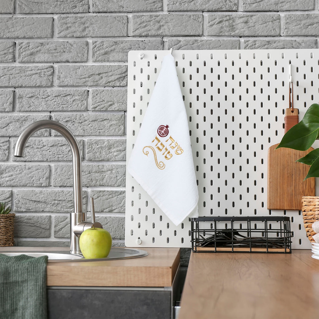 A white kitchen towel with a gold and red coffee cup graphic hangs on a white pegboard in a modern kitchen setting with a gray brick wall, wooden countertop, and a green apple on the sink.