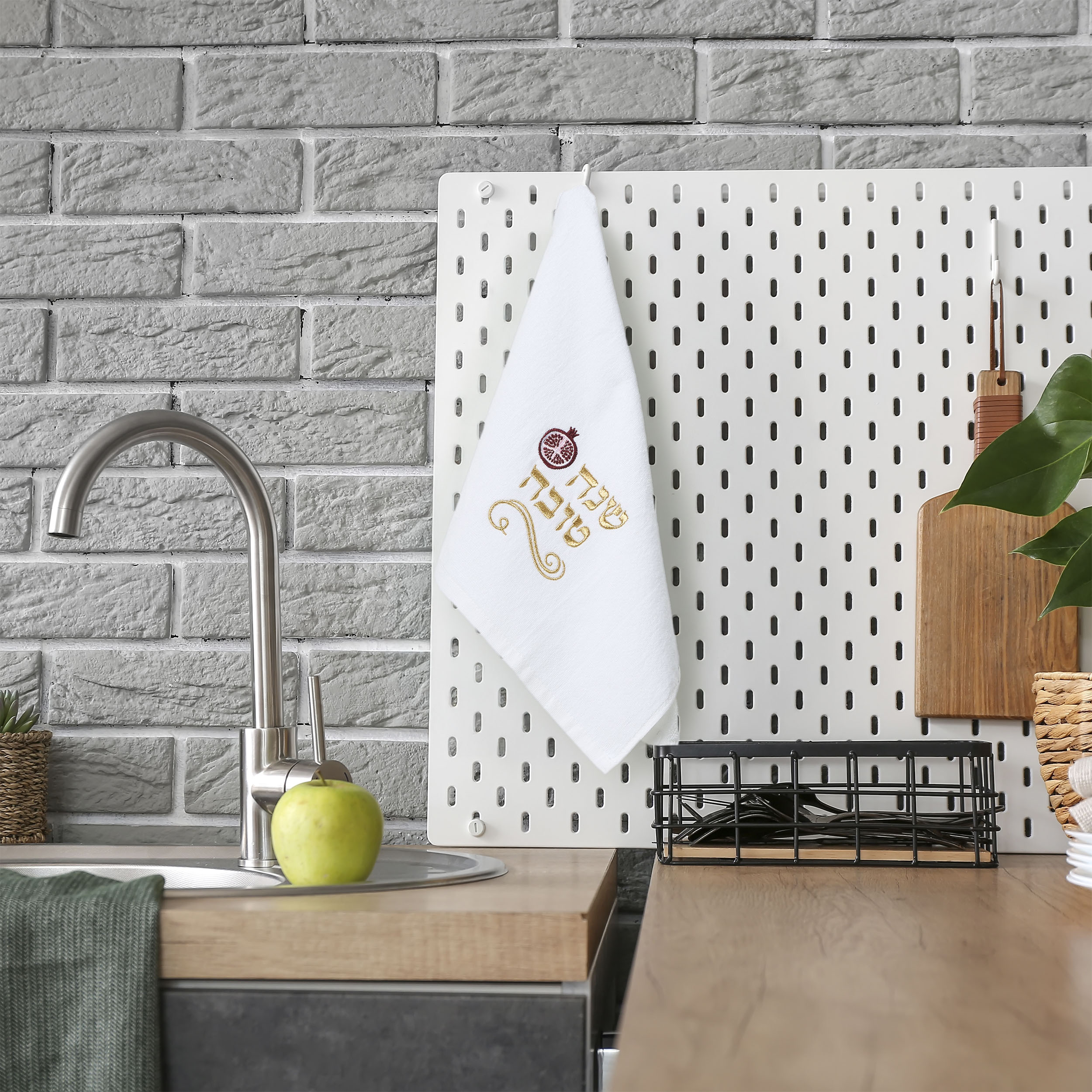 A white kitchen towel with a gold and red coffee cup graphic hangs on a white pegboard in a modern kitchen setting with a gray brick wall, wooden countertop, and a green apple on the sink.