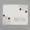 A square white fabric cloth with embroidered floral designs in beige, red pomegranate motifs, and the text 'Shana Tova' in English and Hebrew, displayed on a neutral gray background.