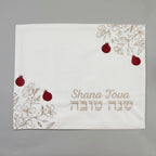 A square white fabric cloth with embroidered floral designs in beige, red pomegranate motifs, and the text 'Shana Tova' in English and Hebrew, displayed on a neutral gray background.