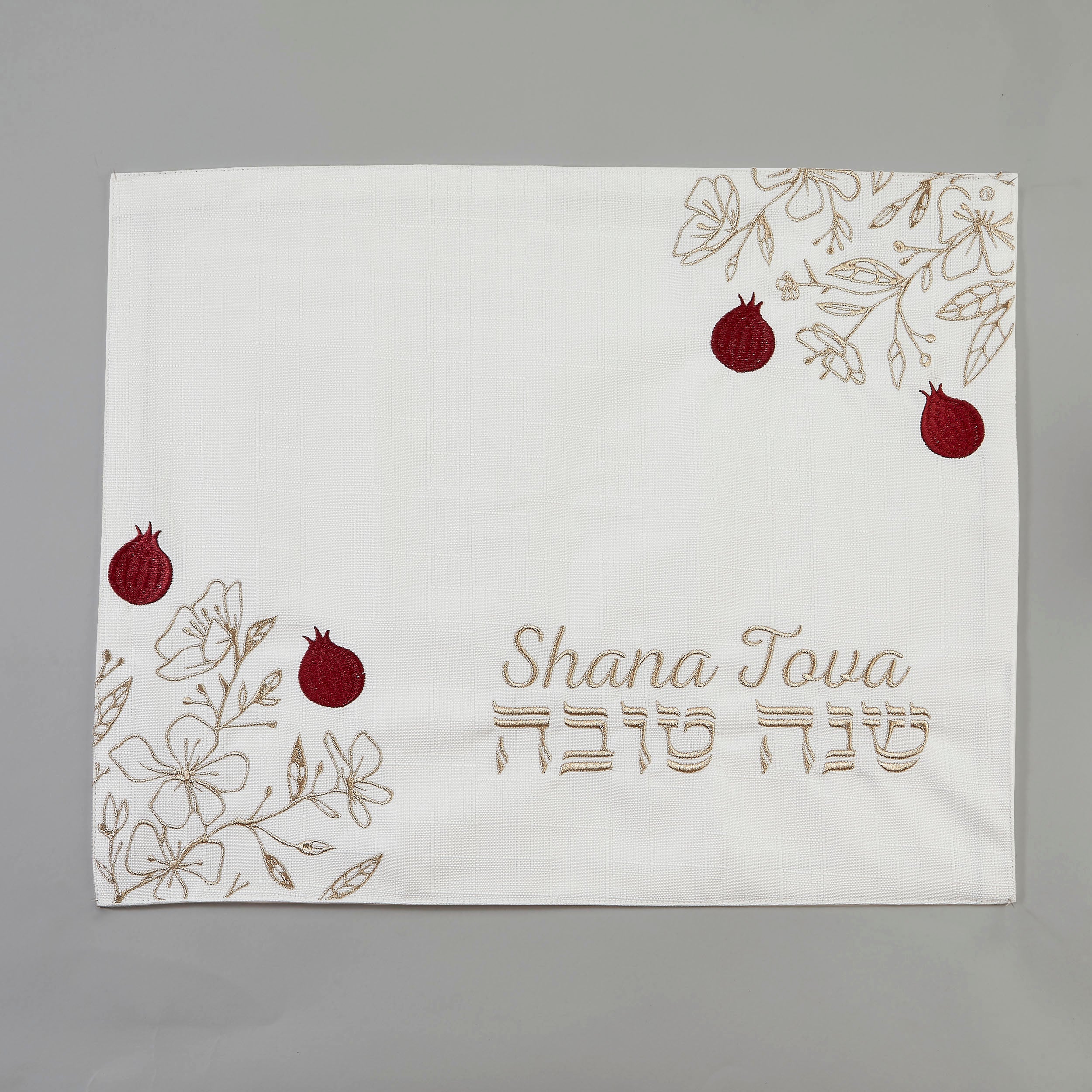 A square white fabric cloth with embroidered floral designs in beige, red pomegranate motifs, and the text 'Shana Tova' in English and Hebrew, displayed on a neutral gray background.