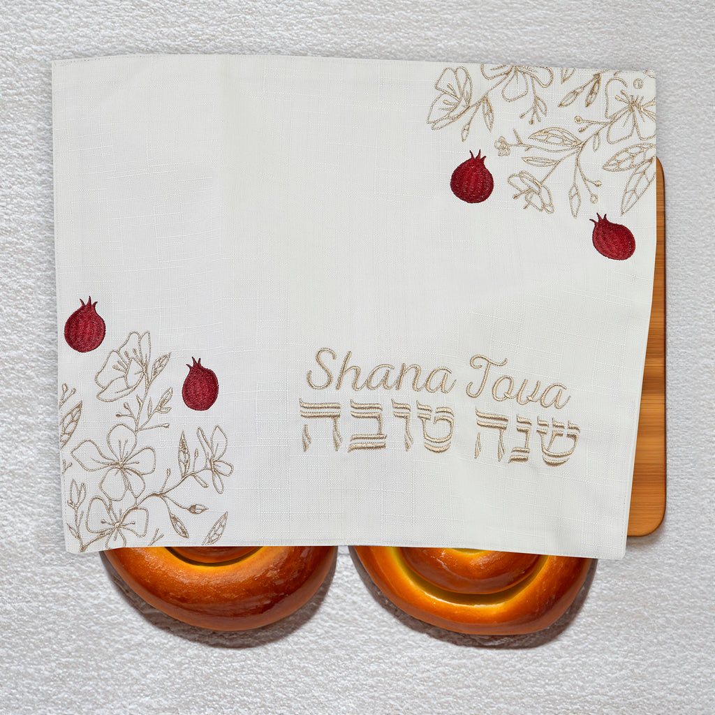 A white fabric challah cover with embroidered floral designs, red pomegranate motifs, and the Hebrew and English text 'Shana Tova' in the center, draped over two round challahs on a white textured wall.