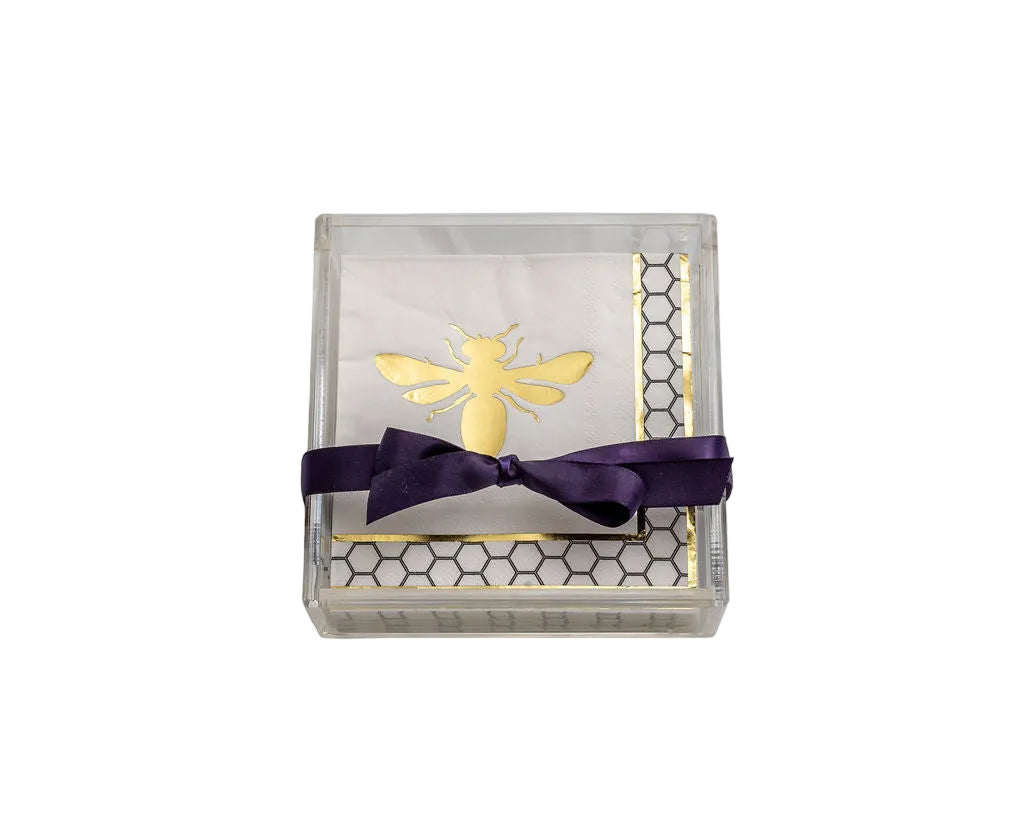 A square clear acrylic gift box containing a white napkin with a gold bee design, a gold honeycomb border, and a dark purple satin ribbon tied in a bow, set against a plain white background.
