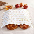 Honeycomb-patterned challah cover with 'Shana Tova' text, apples, and honey dipper on a light background.
