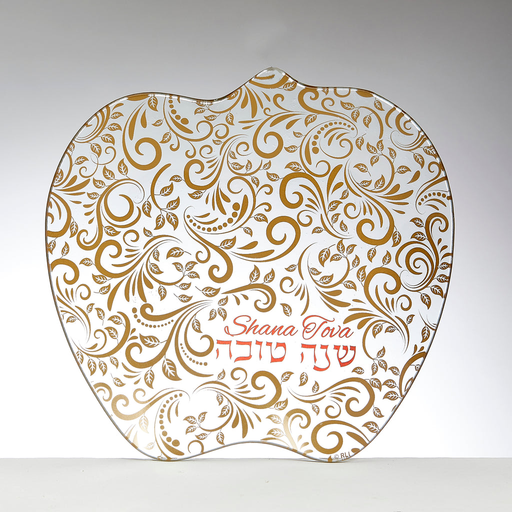 An apple-shaped glass plate with a white background decorated with an intricate gold paisley and leaf pattern. The Hebrew text 'Shana Tova' and the English phrase 'Happy New Year' are written in red cursive in the lower right area.