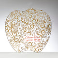 An apple-shaped glass plate with a white background decorated with an intricate gold paisley and leaf pattern. The Hebrew text 'Shana Tova' and the English phrase 'Happy New Year' are written in red cursive in the lower right area.