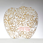 An apple-shaped glass plate with a white background decorated with an intricate gold paisley and leaf pattern. The Hebrew text 'Shana Tova' and the English phrase 'Happy New Year' are written in red cursive in the lower right area.