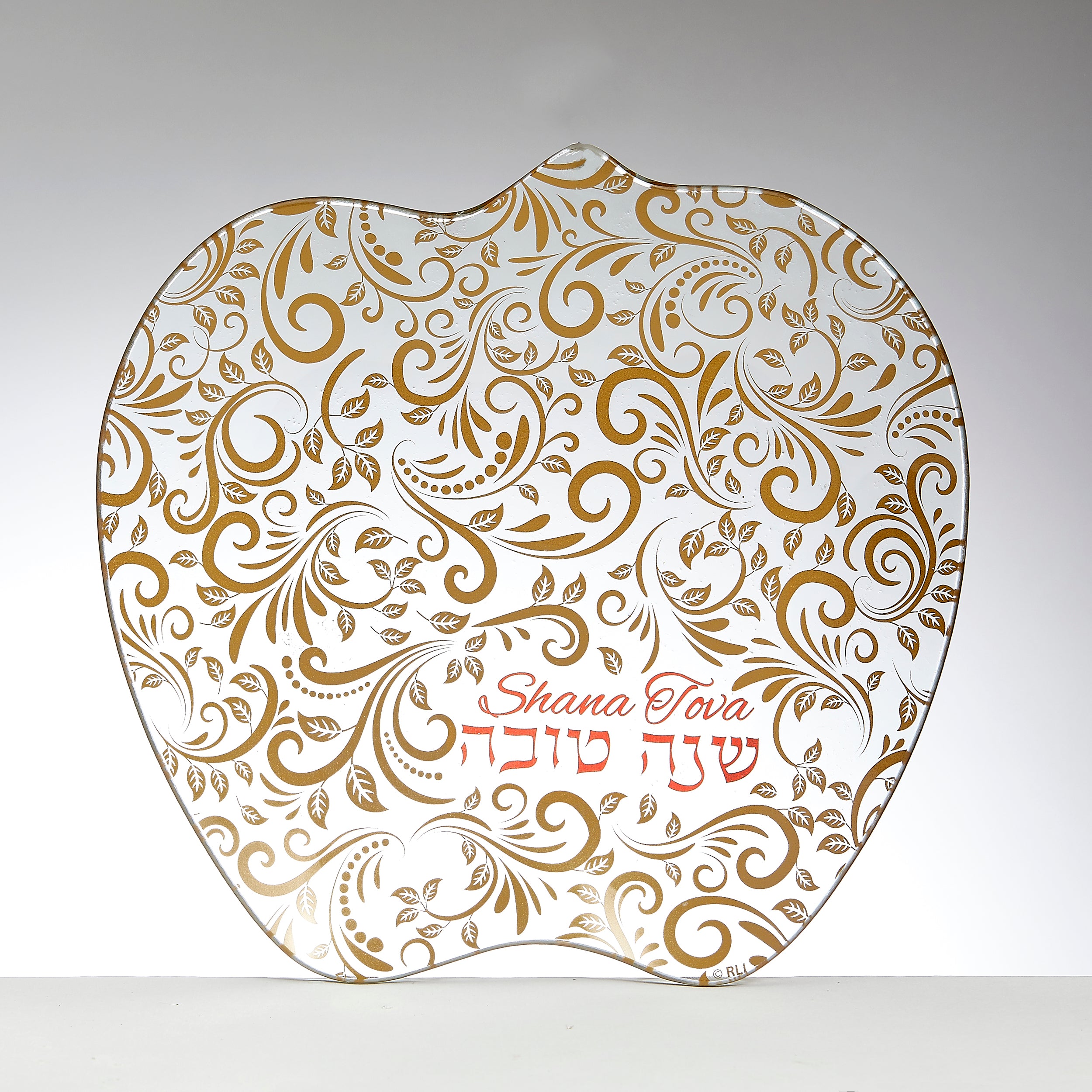 An apple-shaped glass plate with a white background decorated with an intricate gold paisley and leaf pattern. The Hebrew text 'Shana Tova' and the English phrase 'Happy New Year' are written in red cursive in the lower right area.